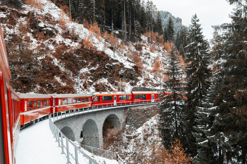 A vibrant red train crosses a snowy coniferous landscape on a scenic mountain bridge in winter.